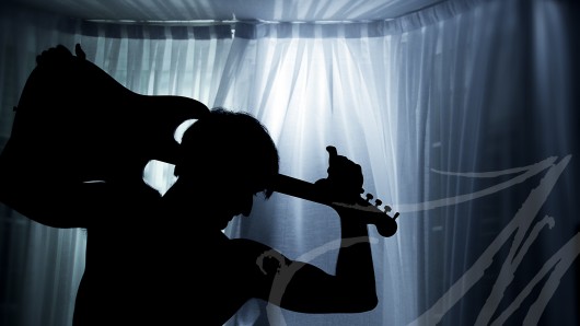 Fotografía de la silueta de un hombre con una guitarra sobre su hombro bajo la luz azul de una ventana, de noche.