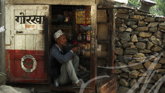 Fotografía social de viajes. Nepal. Hombre mayor con tapi regenta un minúsculo quiosco mientras fuma