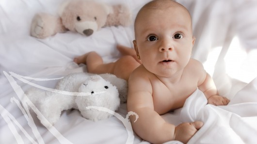 Fotografía de una bebé de cinco meses, sobre la cama, entre sus muñecos